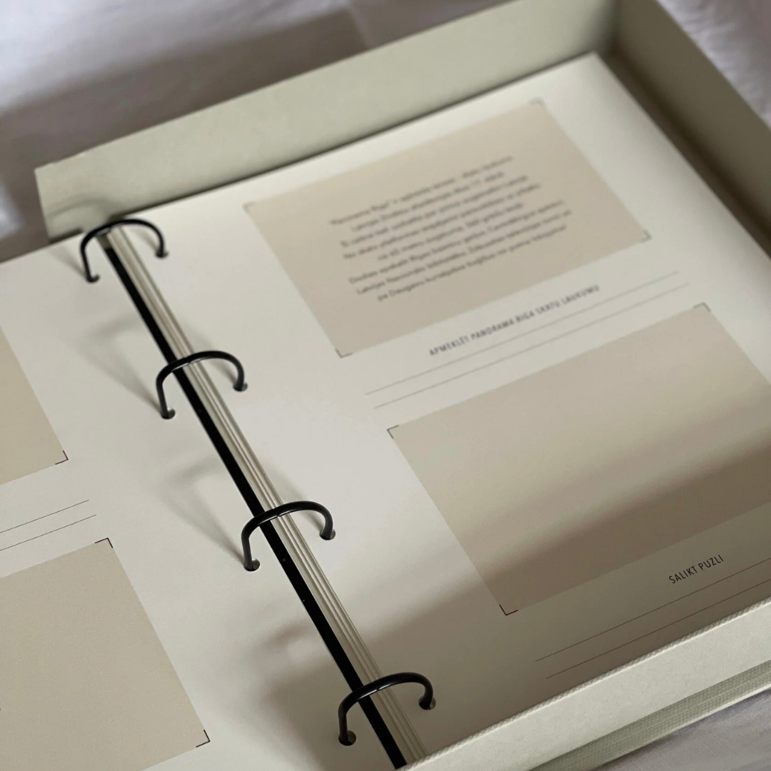 Fotoalbums "Bildē būt" ar bēšu vāku un melnu vāka apdruku. Albumam ir magnētiska aizdare un melns metāla riņku mehānisms iekšlapu stiprināšanai.